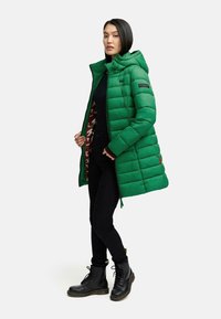 Giacca imbottita verde con cappuccio, chiusura con zip e fodera a motivo mimetico, abbinata a jeans skinny neri e stivali neri con lacci.