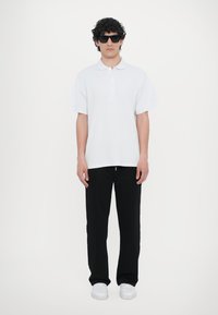 Hvit poloskjorte med korte ermer og tre-knaps knappestolpe, kombinert med sorte bukser og hvite sneakers. Modellen har på seg svarte solbriller.