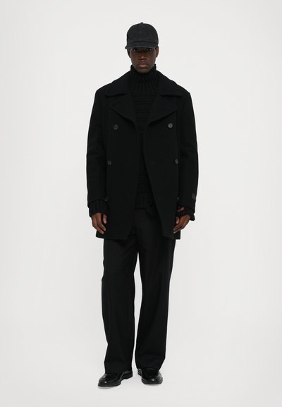 Svart peacoat med dubbelknäppt design, ulltextur, hög krage; lager på lager med en ribbad svart polotröja och löst sittande svarta byxor.