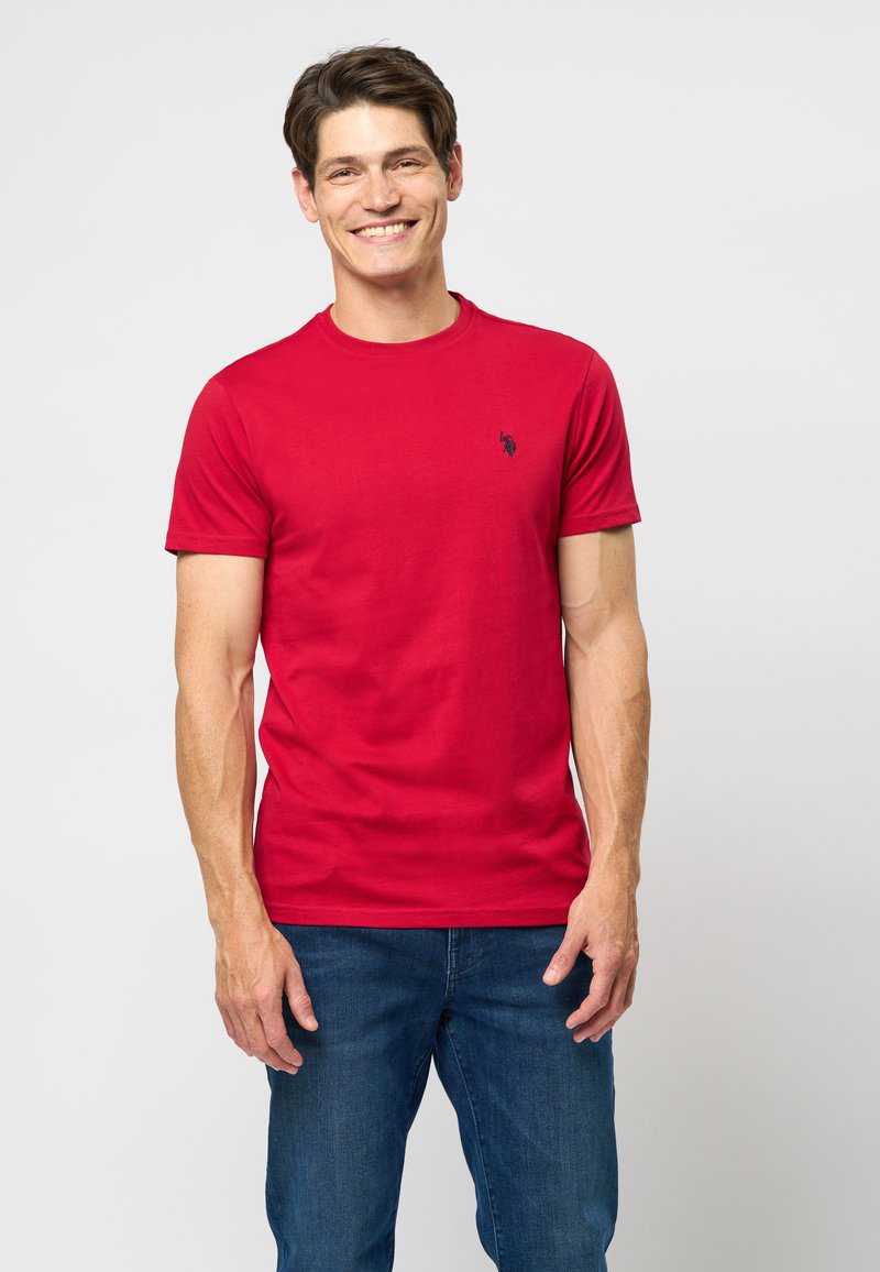 Tricou roșu din bumbac, cu guler rotund și mâneci scurte, având un mic logo negru pe pieptul stâng. Asociat cu blugi din denim albastru.