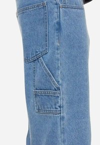 Macacão de ganga em azul claro, com vários bolsos, corte reto e costuras visíveis. Material de algodão durável.