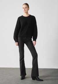 Svart ribbestrikket genser med avslappet passform, kombinert med mørke flarejeans. Modellen har på seg svarte ankelstøvler og står mot en lys bakgrunn.