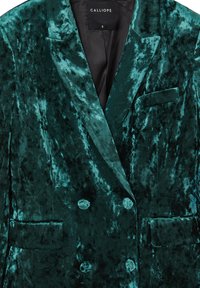 Blazer doppiopetto in velluto verde con fodera nera, revers appuntiti e fazzoletto da taschino. Il tessuto testurizzato mostra una leggera lucentezza con schemi marmorizzati.