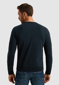 Felpa blu navy con collo a giro e polsini a costine, caratterizzata da una texture liscia e un dettaglio sulla spalla, abbinata a jeans in denim.