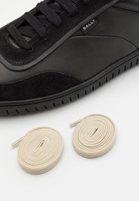 Baskets noires Bally avec des détails en daim et deux paires de lacets beiges enroulés sur une surface blanche.