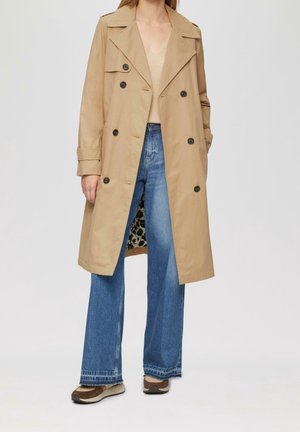 Kobieta nosi beżowy dwurzędowy trencz na beżowym swetrze i szerokich niebieskich dżinsach oraz brązowo-białe buty.