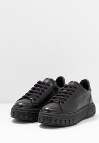 Baskets en cuir noir avec un bout rond, semelle en caoutchouc noir épaisse présentant une texture tressée, et lacets plats noirs. Les détails incluent un petit logo.