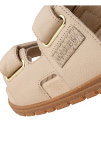 Beige lädersandal med två breda remmar, guldfärgad hårdvaruaccent, texturerade detaljer och en brun gummisula.