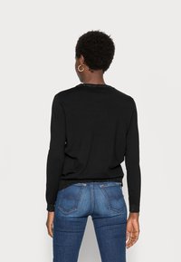 Schwarzer Pullover mit rundem Halsausschnitt, langen Ärmeln und einem strukturierten, glitzernden Saum. Kombiniert mit blauen Jeans, die eine schlanke Passform zeigen.
