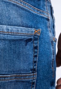 Jeans de denim con un acabado azul, que presentan costuras en contraste y un pequeño detalle en el bolsillo trasero. La tela tiene un aspecto texturizado.