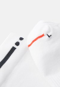 Calcetines blancos con tela texturizada; presentan acentos en negro y naranja, incluyendo una franja y pequeños puntos. La vista de cerca resalta la costura.