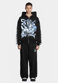 Czarna bluza z zamkiem błyskawicznym z niebieskim nadrukiem graficznym, z grupowym zdjęciem z przodu. W zestawie z dopasowanymi czarnymi spodniami dresowymi i trampkami.