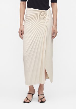 Žena, ktorá nosí krémovú plisovanú midi sukňu so bočným rozparkom, v kombinácii s čiernym topom a čiernymi sandálmi s remienkami a zlatými akcentmi.