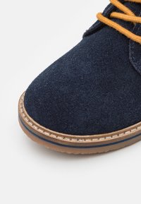 Granatowe zamszowe buty z okrągłym noskiem, zdobione brązowym sznurowadłem oraz przeszyciami wzdłuż brązowej gumowej podeszwy.