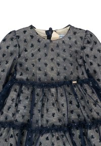 Granatowa sukienka dla małych dziewczynek z długimi przezroczystymi rękawami, pokryta małymi wzorami w kokardy i falbaniastymi warstwami nad beżową sukienką podstawową.
