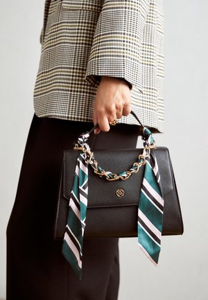 Persona sosteniendo un bolso negro con textura, cadena dorada y bufanda a rayas verde, blanca y negra, vistiendo un blazer a cuadros y pantalones oscuros.