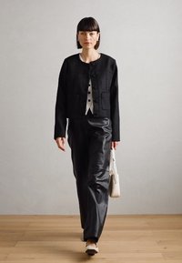 Chaqueta negra texturizada con botones frontales, llevada sobre una camisa clara, combinada con pantalones de cuero negro brillantes y un bolso blanco.