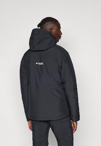 Čierna puffer bunda s kapucňou, vyrobená z odolného materiálu, s dlhšou zadnou časťou a logom na hornej časti chrbta. Navrhnutá pre teplo a pohodlie.