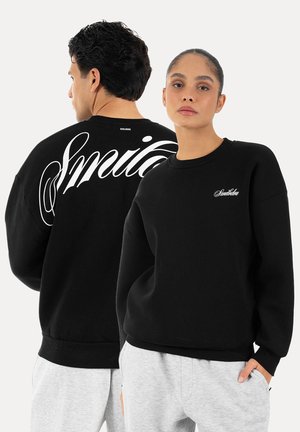 Mężczyzna i kobieta ubrani w czarne bluzy z białym napisem „Smilodox”, mężczyzna tyłem, kobieta przodem, oboje w jasnoszarych spodniach.