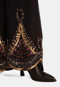 Zwarte kleding met ingewikkelde goud, rood en zilver kraalwerk. Heeft een spitse zwarte hak en toont een gedetailleerd zoompatroon.