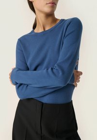 Langarmblaues Sweatshirt aus weichem Strickstoff, mit einem runden Halsausschnitt und gerippten Manschetten, kombiniert mit schwarzen, hoch geschnittenen Hosen.