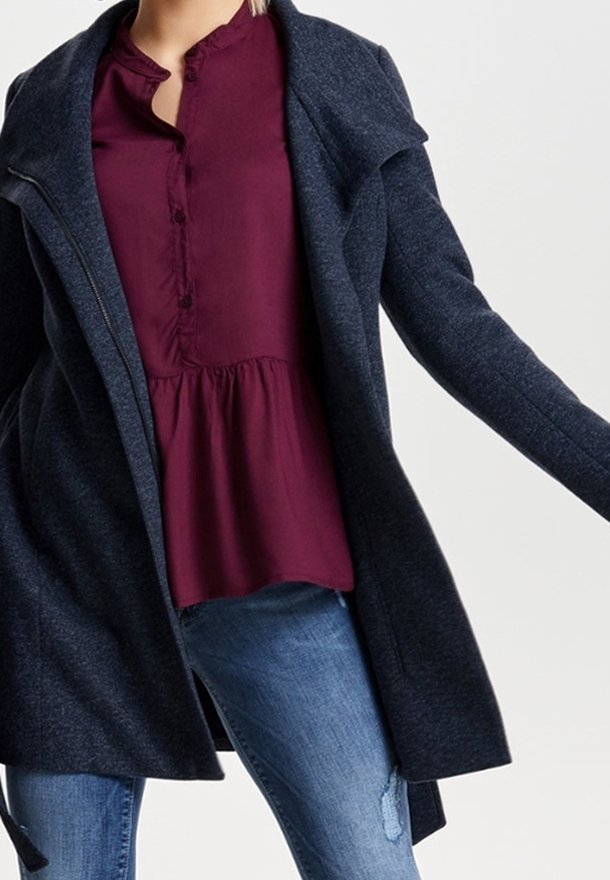 Cappotto blu navy con finitura testurizzata, ampi revers, apertura frontale e zip nascosta. Indossato sopra una camicetta bordeaux e jeans strappati.