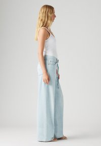 Lyseblå vide denimjeans med høy livhøyde og knytebelte, med en falmet tekstur og rette ben. Modell med en hvit topp.