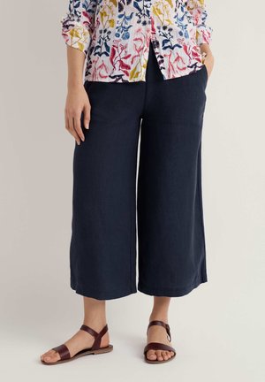 Woman wearing navy wide-leg cropped pants, floral button-up shirt, and brown flat sandals standing with hands in pockets.