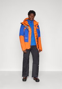 Χρωματιστή μόνωτη ζακέτα σε μπλε και πορτοκαλί χρώμα με λαμπερή υφή, φορεμένη πάνω από μια μπλε ζακέτα, συνδυασμένη με σκούρους παντελόνες και μαύρα παπούτσια.