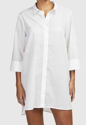 Vrouw die een loszittende witte overhemdjurk draagt met driekwart mouwen en een kraag, tegen een neutrale achtergrond.