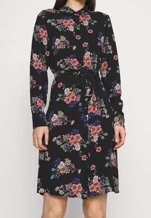 Schwarzes, langärmeliges Kleid mit Knöpfen, rosa, blauem und weißem Blumenmuster und gebundenem Taillengürtel, getragen von einer stehenden Person.