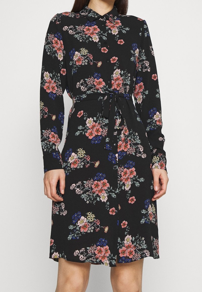 Robe noire à manches longues boutonnée, avec imprimé floral rose, bleu et blanc, et ceinture nouée à la taille, portée par une personne debout.