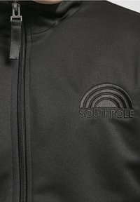 Veste zippée noire en tissu lisse, ornée d'un design arc-en-ciel en relief et du texte "SOUTHPole" sur le côté gauche.