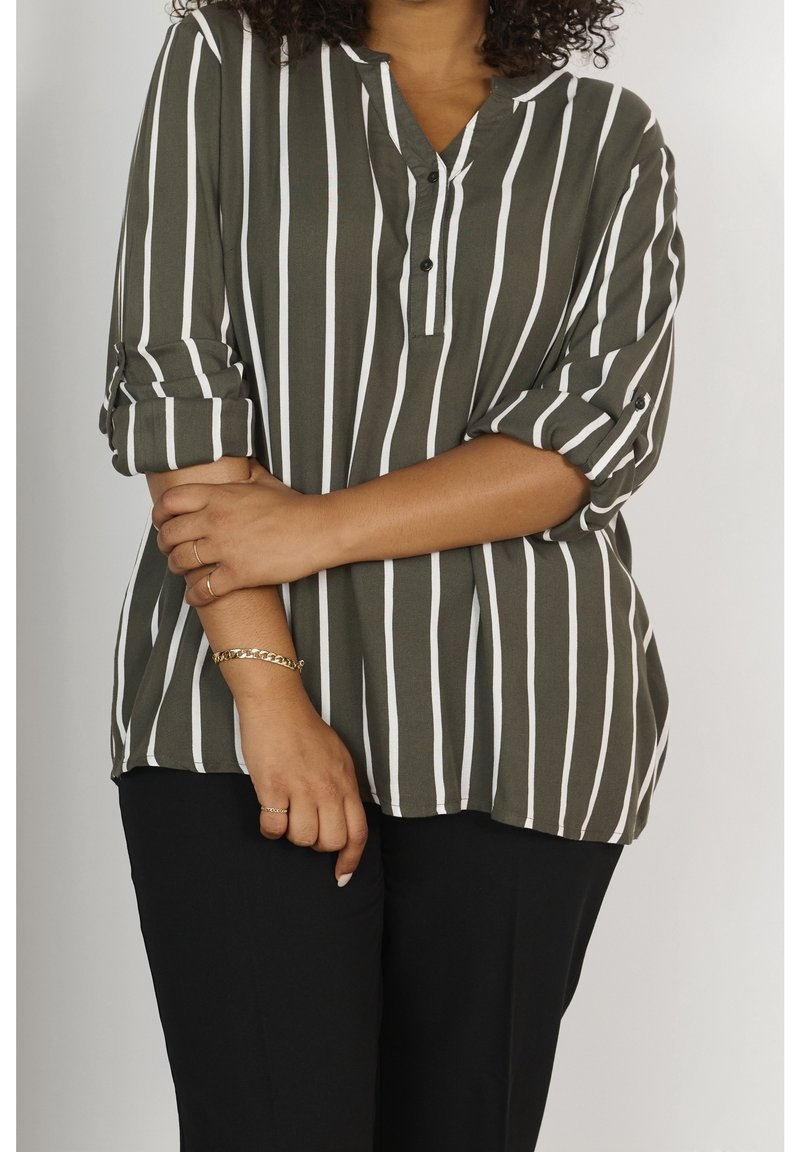 Chemise vert olive à rayures verticales blanches, col à revers, patte de boutonnage et manches retroussées. Portée avec un pantalon noir.