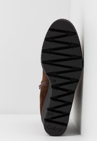 Semelle d'une chaussure en suède marron avec motif de traction en zigzag, fermeture éclair latérale, et nom de marque "Gabor" sur le talon dans un coin blanc.