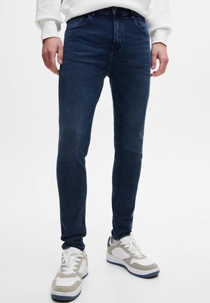 Homme portant un jean bleu foncé slim et des baskets blanches, debout devant un fond blanc uni.