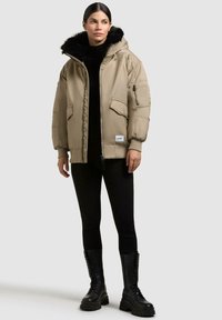 Giacca puffer beige con cappuccio foderato in pelliccia, zip e dettagli sulle tasche, polsini a coste e una vestibilità morbida, abbinata a pantaloni neri e stivali da combattimento.