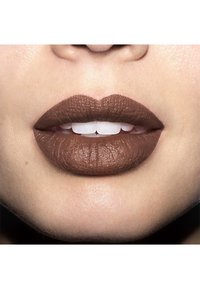 Rossetto marrone opaco sulle labbra che mostra una texture liscia; la luce si riflette sulla pelle leggermente secca che circonda la bocca. Denti bianchi visibili.