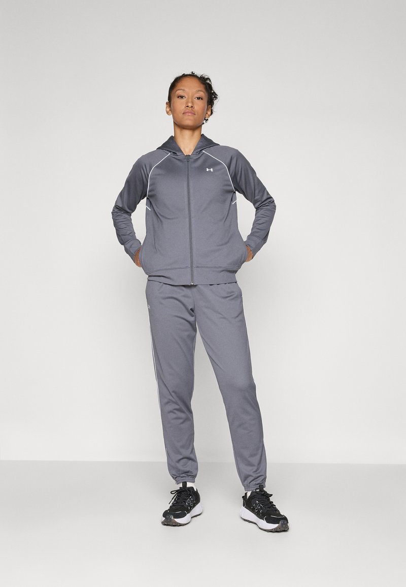 Grå idrettsdrakt laget av lett stoff, med en hettegenser med glidelås og hvite detaljer, samt matchende tapered bukser. Svarte joggesko fullfører antrekket.