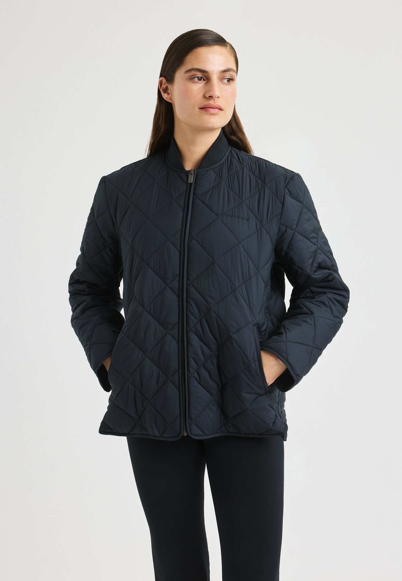 Kvinna iklädd en svart vadderad zip-up jacka med händerna i fickorna, som ser något åt höger, stående mot en ljus, enhetlig bakgrund.