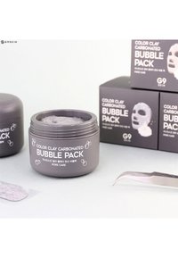 Pot gris étiqueté "Pack de Bulles Carbonat�ées Argile Colorée" avec des illustrations de bulles, accompagné de boîtes en carton gris et d'un outil de pincette pour l'application.