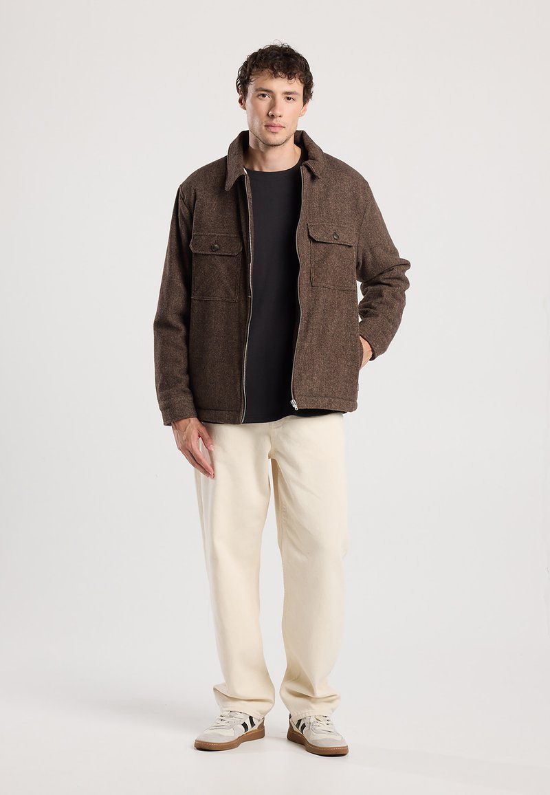 Bruine wollen jas met een ritssluiting aan de voorkant, twee borstzakken en een kraag, gecombineerd met een zwart shirt en crèmekleurige broek. Casual schoenen.
