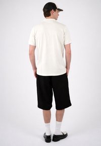 Homme portant un polo blanc, un short noir à hauteur des genoux, des chaussettes blanches, des chaussures noires et une casquette noir et blanc, debout de dos sur un fond uni.