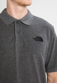 Grå pikétröja i strukturväv, med en svart broderad logotyp på vänster bröst och en klassisk krage med två knappar.