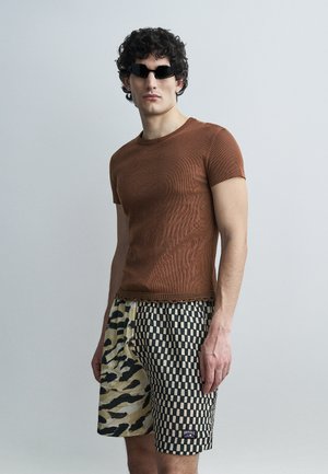 Jeune homme portant une chemise marron à manches courtes, des lunettes de soleil noires et un short patchwork avec des motifs damier et imprimé animal.