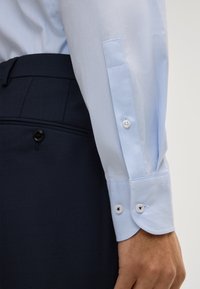 Camicia di stoffa azzurro chiaro con texture e polsini abbottonati, abbinata a pantaloni navy con texture e chiusura a un bottone.
