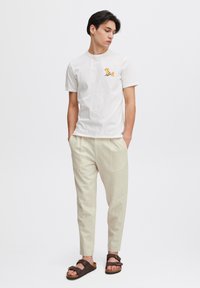 Cremfarbenes T-Shirt mit einem kleinen gelben und orangefarbenen Grafik, kombiniert mit hellbeigen, lässig geschnittenen Hosen und braunen Sandalen.