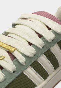 Κοντινό πλάνο κορδονιών αθλητικού παπουτσιού πάνω σε ροζ γλώσσα, πράσινη πλευρά από σουέτ με λευκές ραβδωτές ρίγες και μεταλλική χρυσή ετικέτα με το λογότυπο.