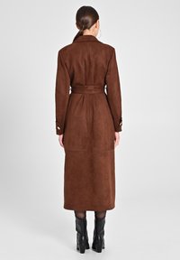 Brauner Wildleder-Trenchcoat mit tailliertem Schnitt, langen Ärmeln und mittellanger Länge, kombiniert mit schwarzen Ankle Boots. Glatte Textur, minimale Details.