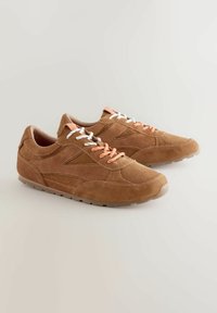 Braune Wildledersportschuhe mit niedrigem Profil, versehen mit weißen und orangenen Schnürsenkeln, strukturierten Details und einer Gummisohle.